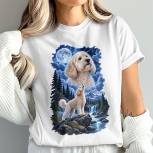 May include: A white short-sleeved t-shirt featuring a graphic print of a golden-colored dog. The design includes a portrait of the dog above a full-body image of the dog standing on a rock near a waterfall, pine trees, and a full moon.