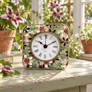 May include: A square table clock featuring a white face with black Roman numerals and black hands. The frame is constructed from a gold-toned metal lattice decorated with multi-coloured floral cutouts in shades of red, green, and blue.