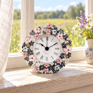 May include: A round quartz clock featuring a white face with black Roman numerals and hands. The frame is decorated with a dense arrangement of small, multi-coloured enamelled flowers in shades of pink, white, and dark blue, accented with tiny crystals.