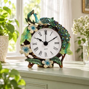May include: A decorative quartz table clock featuring a sculpted peacock with blue rhinestone accents, surrounded by green leaves and light blue flowers with central crystals. The white clock face displays black Roman numerals and hands.