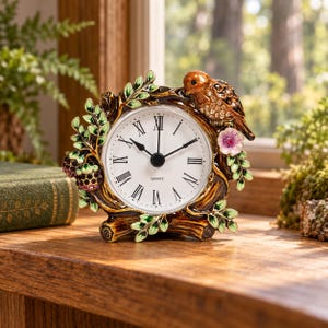 May include: A round quartz clock with a white face and black Roman numerals, encased in a decorative frame shaped like a tree branch. The frame features a brown bird with clear crystals, green leaves, a pink flower, and dark purple berry clusters.