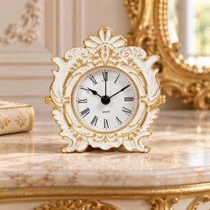 Könnte beinhalten: Eine weiß-goldene Tischuhr mit einem verzierten, geschwungenen Rahmen und einem runden Zifferblatt. Die Uhr hat schwarze römische Ziffern, schwarze Stunden- und Minutenzeiger sowie das Wort QUARTZ in schwarzer Schrift auf dem weißen Zifferblatt.