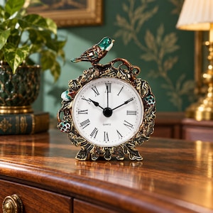 May include: A round quartz clock featuring a white face with black Roman numerals and hands. The clock is encased in a decorative metal frame shaped like leaves and berries, topped with a small, colourful bird figure with green and brown enamel.