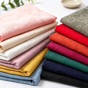 Könnte beinhalten: Eine Sammlung gefalteter Stoffquadrate in verschiedenen Unifarben wie Rosa, Creme, Weiß, Mauve, Gelb, Hellblau, Olivgrün, Rot, Orange, Magenta, Dunkelgrün, Marineblau und Schwarz, die alle ein strukturiertes Gittermuster aufweisen.