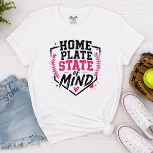 May include: A white short-sleeve t-shirt featuring a black and pink graphic of a home plate shape with baseball stitching and the text "HOME PLATE STATE of MIND" with a small pink heart. The shirt has a crew neckline and a standard fit.