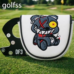 May include: A white golf putter headcover featuring an embroidered dark grey bear with a stitched mouth, a yellow button eye, and red accents. The black metal putter base is visible behind the cover, displaying the text DF3 and the word golfss.