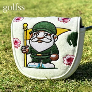 May include: A white golf putter headcover featuring an embroidered gnome wearing a green outfit and hat, holding a yellow flag. The cover is decorated with pink hibiscus flowers and the text golfss in the upper left corner.