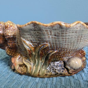 May include: A ceramic bowl shaped like a large seashell with a glossy finish. The exterior features a textured grid pattern in shades of brown and cream, accented with sculpted green seaweed and small shell details along the base rim.