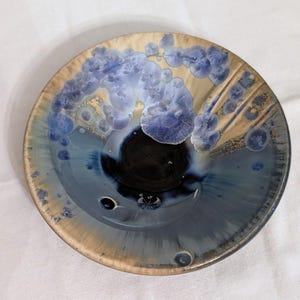 May include: A round ceramic bowl featuring a crystalline glaze with a deep black center that transitions into shades of blue and tan. The surface displays distinct, flower-like crystal formations in light blue, scattered across the rim and interior.