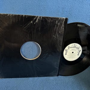 May include: A black vinyl record with a white label featuring the text "Grand Royal" and a black record sleeve. The record is partially out of its sleeve, revealing the shiny black surface. The sleeve has a circular cutout.