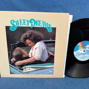 May include: Vintage vinyl record album for the movie soundtrack "Sweet Dreams." The album cover features a couple in a car with the title in teal script. A black vinyl record is next to the album cover.