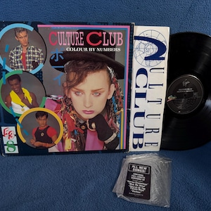 Op de afbeelding: Een Culture Club Colour by Numbers vinyl platen set met een vierkante albumhoes met een portret van Boy George en drie cirkelvormige inzetstukken van bandleden, een witte binnenhoes met blauwe tekst, een zwarte vinylplaat en een plastic zakje.