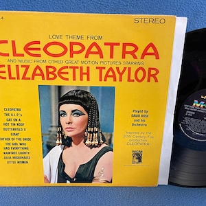 May include: A yellow and red vinyl record album cover with a photo of Elizabeth Taylor as Cleopatra. The album title is "Love Theme From Cleopatra" and includes music from other films starring Elizabeth Taylor. The album is by MGM Records.
