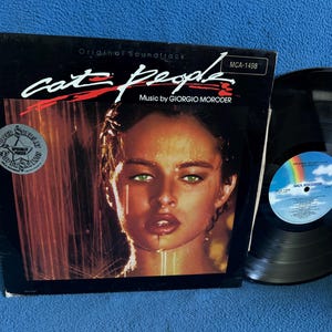 May include: Vinyl record album for the movie "Cat People" with a woman's face on the cover. The album cover is black with the title in white and red. The record is black with a blue and rainbow label.