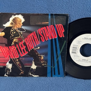 May include: A black vinyl record with a red and white label. The label features the text "David Lee Roth" and "Stand Up". The record is a 45 rpm single.