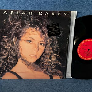 May include: Vinyl record album cover for Mariah Carey's debut album. The album cover features a portrait of Mariah Carey with curly hair. The record is black with a red and white label. The album title is "Mariah Carey".