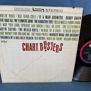 May include: Vintage vinyl record album titled "Chart Busters" with a white and grid-patterned cover. The cover lists song titles and artist names in green and black text. A black vinyl record is visible next to the album.