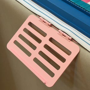May include: A light pink plastic shelf with a slotted surface and two hinges, attached to the edge of a blue and white striped object. The shelf features a rectangular design with horizontal cutouts and a small lip at the bottom edge.