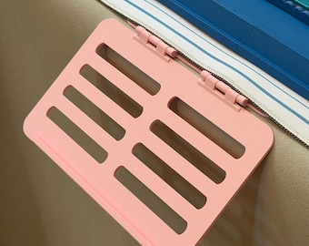 May include: A light pink plastic shelf with a slotted surface and two hinges, attached to the edge of a blue and white striped object. The shelf features a rectangular design with horizontal cutouts and a small lip at the bottom edge.
