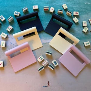 May include: A collection of six mahjong tile racks in navy blue, cream, and light purple, arranged on a surface with scattered mahjong tiles featuring various colorful symbols, characters, and patterns in red, green, and blue ink.