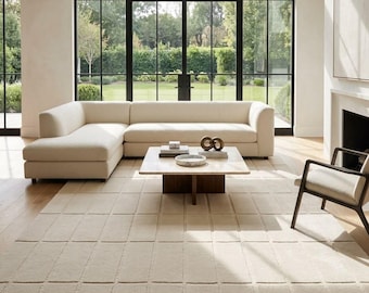 May include: A large, cream-coloured sectional sofa with a chaise lounge, positioned on a textured, grid-patterned area rug. A square coffee table with a light stone top and dark wooden base sits in front of the sofa, holding books and a decorative object.
