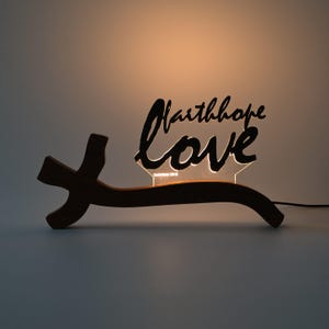 Könnte beinhalten: Ein dunkler, kreuzförmiger Holzsockel trägt ein hinterleuchtetes Acrylschild mit den Wörtern faith, hope und love in einer schwarzen Schreibschrift. Der Text wird von hinten beleuchtet und Corinthians 13:13 ist darunter zu sehen.