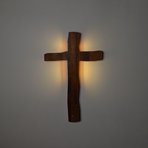 May include: A dark brown wooden cross with a natural, uneven edge texture. The cross is illuminated from behind, casting a soft, warm golden glow onto the surrounding area, highlighting the vertical and horizontal grain of the wood surface.
