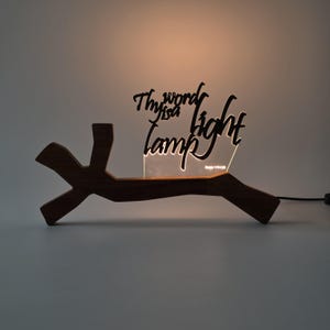 Könnte beinhalten: Ein dunkelbrauner Astsockel aus Holz trägt eine von unten beleuchtete, klare Acrylplatte. Die Platte zeigt den schwarzen Schriftzug Thy word is a lamp unto my feet and a light unto my path sowie die Quellenangabe Psalm 119:105 am unteren Rand.