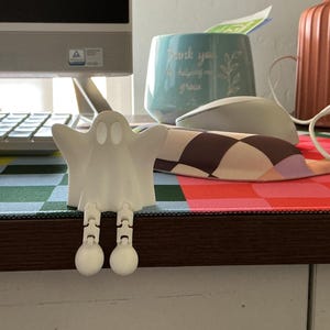 May include: A small, white, 3D-printed ghost figurine with articulated, dangling legs. The ghost features a smooth, matte finish, hollow oval eyes, and a rounded head, designed to sit on the edge of a desk or shelf with its legs hanging down.