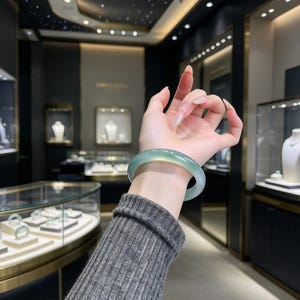 Puede incluir: Un brazalete de jade verde claro, translúcido y liso, con un acabado pulido. El brazalete circular presenta variaciones sutiles en la intensidad del color, que van desde el verde menta pálido hasta un verde espuma de mar más profundo.