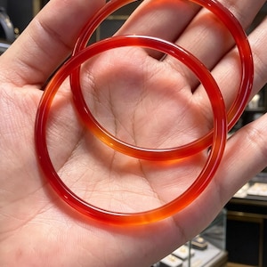 May include: Two smooth, circular bangles crafted from translucent, deep red carnelian stone. The bracelets feature a polished, glossy finish that catches the light, showcasing the natural color variations and clarity of the vibrant red gemstone material.