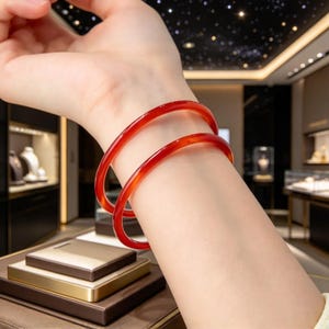 Op de afbeelding: Twee gladde, doorschijnende rode armbanden met een gepolijste afwerking. De ronde armbanden worden samen om de pols gedragen en tonen een levendige, dieprode kleur en een eenvoudig, afgerond profiel dat het licht over het oppervlak weerkaatst.