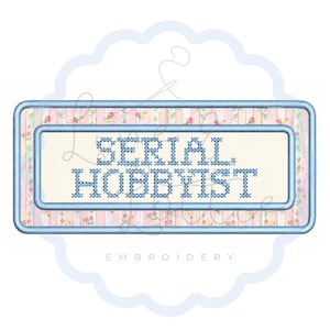 May include: A rectangular embroidered patch featuring a light pink background with a delicate floral pattern. The center displays the text SERIAL HOBBYIST in blue cross-stitch lettering, framed by a double blue stitched border.