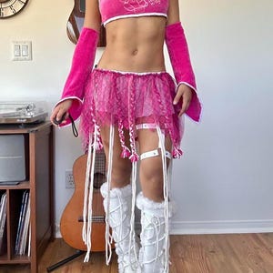 May include: A bright pink Hello Kitty crop top with white trim, matching pink arm warmers, and a layered pink mesh mini skirt featuring hanging braided fringe details. The outfit is paired with white lace-up platform boots with faux fur trim.