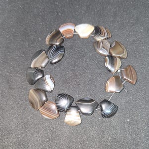 Può includere: Una disposizione circolare di perline in agata a bande lucidate a forma di ventaglio, infilate su un filo sottile. Ogni perlina mostra strati naturali unici di marrone, grigio, nero e bianco, creando un motivo a righe distinto sulle superfici.