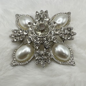 May include: A silver-toned brooch featuring a central large clear rhinestone surrounded by smaller clear rhinestones and four oval-shaped white faux pearls. The metal frame is encrusted with numerous tiny clear rhinestones in a floral pattern.