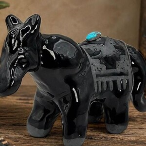 May include: A glossy black ceramic horse figurine featuring a matte gray saddle blanket with a geometric pattern and a single oval turquoise stone set on its back. The horse has four legs with matte gray hooves and a smooth, reflective finish.