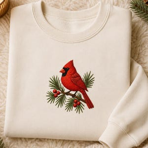 May include: A cream-coloured crewneck sweatshirt featuring an embroidered design of a bright red cardinal bird perched on a green pine branch with small red berries. The embroidery is centred on the chest of the long-sleeved pullover garment.