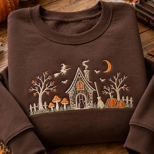 May include: A dark brown crewneck sweatshirt featuring an embroidered scene of a house with a chimney, a witch on a broomstick, a crescent moon, bats, trees, mushrooms, and pumpkins. The design includes a cat silhouette in the doorway.