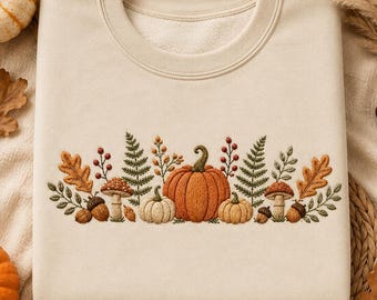 May include: A cream-colored crewneck sweatshirt featuring a central horizontal embroidery design of orange pumpkins, brown acorns, red berries, green fern fronds, and tan mushrooms arranged in a detailed botanical pattern across the chest.
