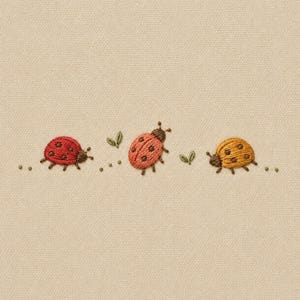 May include: An embroidery design on a beige fabric background featuring three ladybugs in a horizontal row. The ladybugs are colored red, coral, and yellow, each with dark brown spots, antennae, and legs, accompanied by small green leaves.