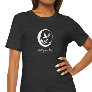 May include: A dark charcoal gray short-sleeve t-shirt featuring a white circular graphic on the chest. The design depicts a dove carrying an olive branch inside a crescent moon shape, with the text "peace grows here" printed below the graphic.