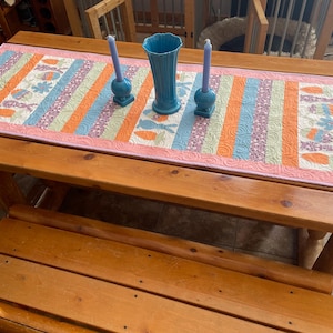 Könnte beinhalten: Ein Holztisch mit passender Bank, bedeckt mit einem farbenfrohen, gesteppten Tischläufer. Der Läufer zeigt vertikale Streifen in Orange, Blau und Lila mit Blumen- und Blättermustern. Zwei blaue Kerzenhalter und eine türkisfarbene Vase stehen auf dem Tisch.