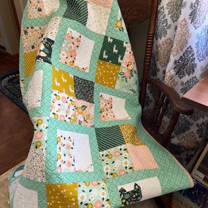 Könnte beinhalten: Eine Patchwork-Decke mit verschiedenen floralen und geometrischen Mustern in Grün-, Gelb-, Rosa- und Weißtönen. Die Decke ist gefaltet und über einen Holz-Schaukelstuhl gelegt.
