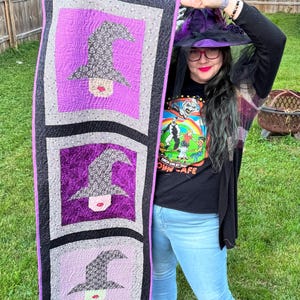 Puede incluir: Un tapiz de pared acolchado con tres paneles con diseños de sombreros de bruja. Cada panel tiene un fondo morado con un sombrero de bruja gris y negro. El edredón está bordeado con tela negra y gris. El tema es Halloween.