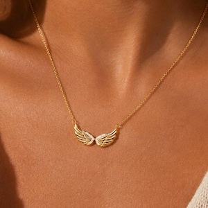 May include: A gold-toned necklace featuring a delicate chain and a central pendant shaped like a pair of symmetrical angel wings. The wings are textured with fine lines to mimic feathers and are accented with small, clear, sparkling stones.
