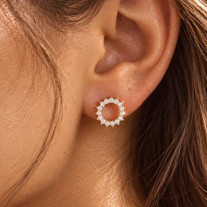 May include: A circular stud earring featuring a gold-toned metal frame set with a series of small, clear, round-cut stones arranged in a sunburst pattern. The earring is positioned on an earlobe, showcasing the reflective quality of the stones.