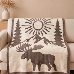 May include: A rectangular crochet blanket featuring a cream-colored base with a dark brown pattern. The design depicts a large moose in the foreground, with mountains, two tall pine trees, and a radiant sun positioned in the upper section.