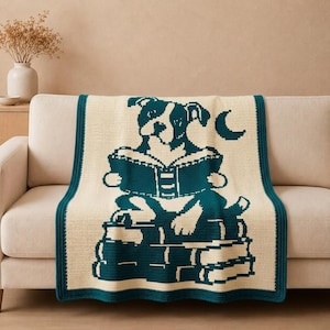 May include: A cream-colored throw blanket featuring a dark teal graphic design of a dog reading an open book while sitting atop a stack of three books, with a crescent moon symbol to the right and a solid dark teal border around the edges.