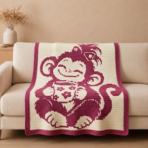 Puede incluir: Una manta de ganchillo rectangular con un diseño bicolor en crema y magenta oscuro. El centro muestra un mono grande y sonriente que sostiene una taza decorada con pequeños patrones de flores. La manta tiene un borde magenta sólido.